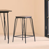 A black metal stool with a simple design featuring a square elm wood seat, supported by slender, angled legs. The stool is positioned next to a matching table against a light-colored wall, creating a modern and minimalist aesthetic