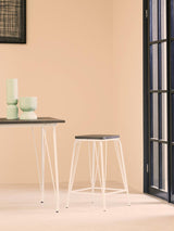 A white metal stool with a natural elm wood seat positioned next to a small table, set against a light-colored wall. The scene includes decorative items on the table and a large window with black frames in the background