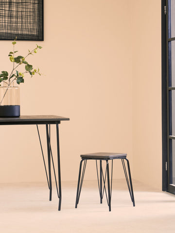 A small black metal stool with a wooden elm seat is positioned next to a sleek black table in a softly lit room with beige walls. A potted plant sits on the table, adding a touch of greenery to the minimalist decor. Natural light streams in through a large window, enhancing the warm atmosphere