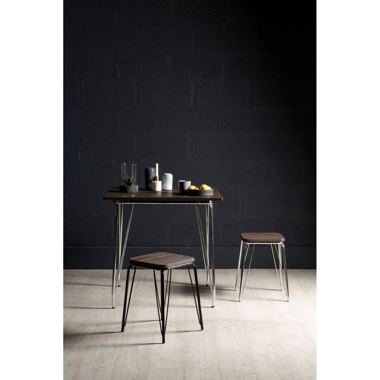 A small black metal stool with a wooden seat made of elm, positioned next to a matching table. The background features a dark wall, and the floor is light-colored wood. On the table, there are glasses, a bottle, and a few pieces of fruit