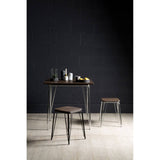 A small black metal stool with a wooden seat made of elm, positioned next to a matching table. The background features a dark wall, and the floor is light-colored wood. On the table, there are glasses, a bottle, and a few pieces of fruit
