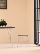 A small wood stool featuring a grey metal frame and a natural elm seat, positioned next to a sleek table in a minimalist room with light-colored walls and a large window. A small potted plant sits on the table, adding a touch of greenery to the scene