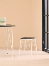 Small wooden stool with a dark elm seat and white metal legs, placed on a light-colored floor. A small table with a dark top and a green decorative item sits nearby against a soft beige wall. Natural light filters in through a window, creating a warm atmosphere