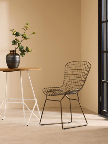Bronze metal grid frame wire chair positioned next to a small wooden table with a vase of flowers, set against a light beige wall and large window