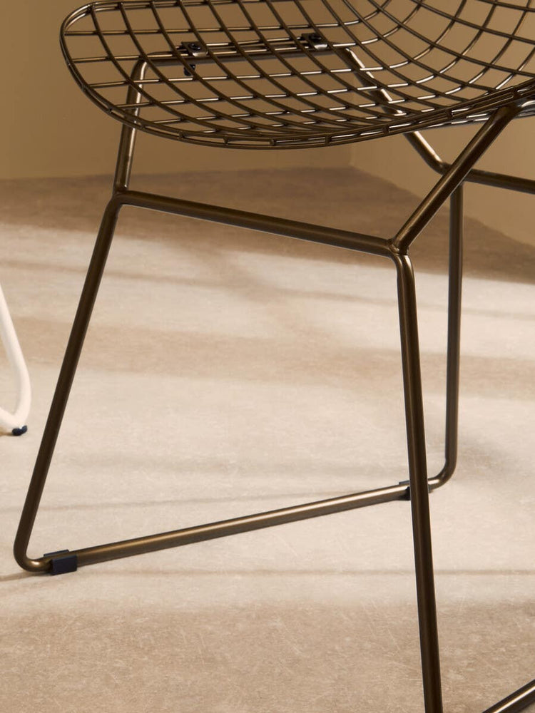 Bronze metal grid frame wire chair with a rounded seat and slender legs, positioned on a light-colored floor. The chair features a modern design with a geometric pattern in the seat and backrest