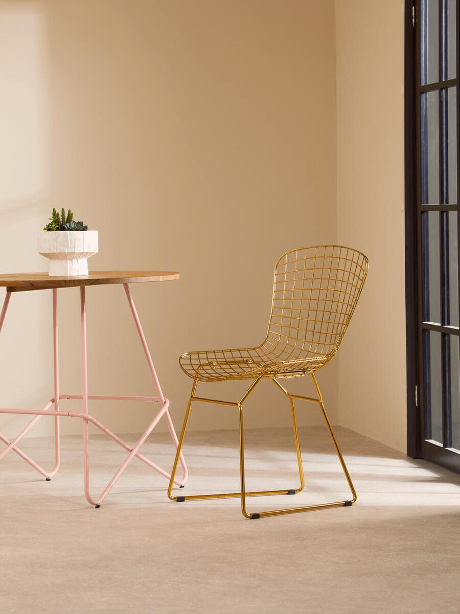 Gold metal grid frame wire chair positioned next to a small round table with a potted plant on top, set against a light beige wall and floor. Natural light streams in from a large window, creating a warm and inviting atmosphere