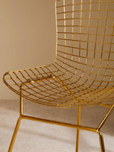 A close-up view of a gold metal grid frame wire chair, showcasing its sleek, modern design with a curved seat and backrest made of thin metal wires, supported by a minimalist base. The background features a soft, neutral wall color that complements the chair's metallic finish