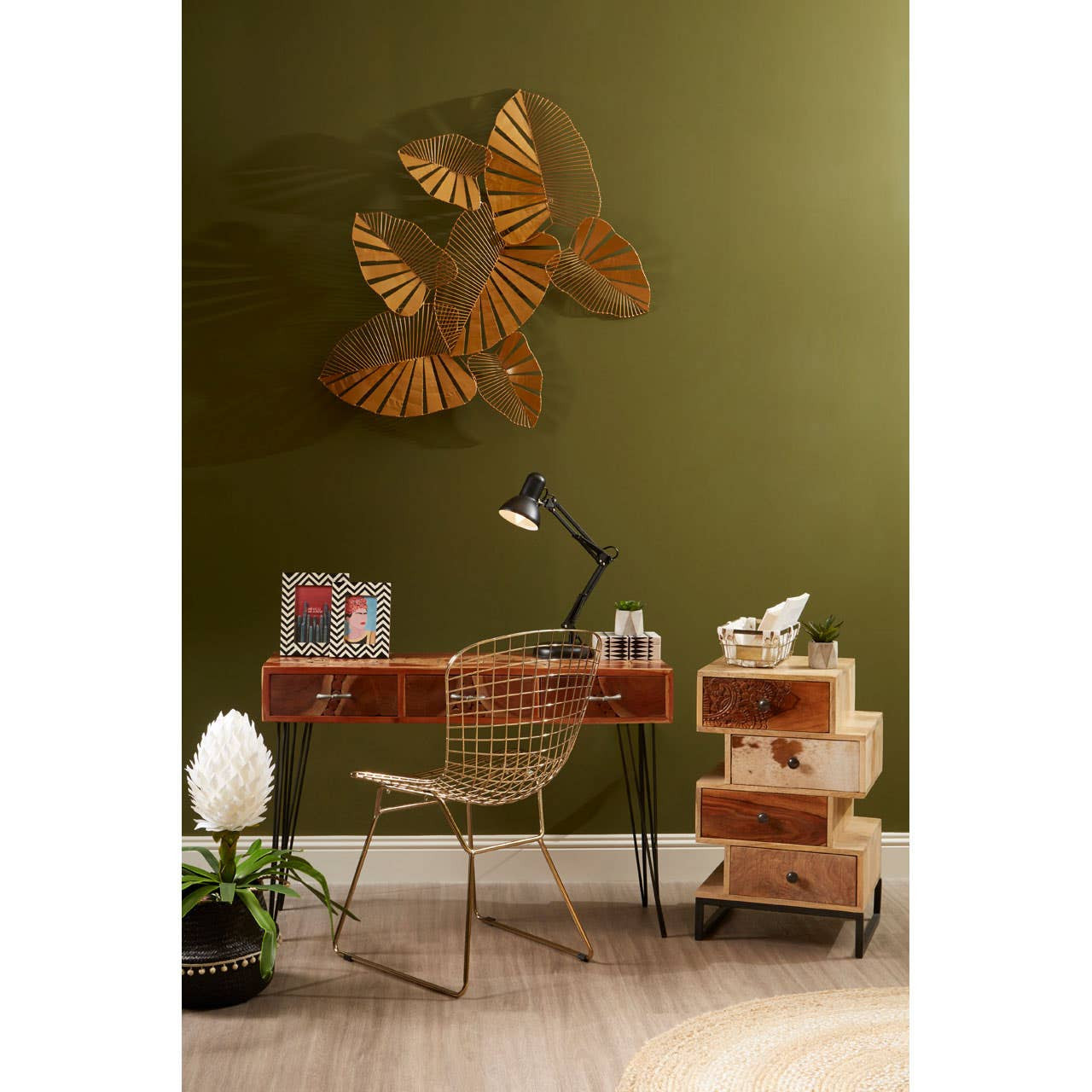 A stylish gold metal grid frame wire chair positioned next to a wooden desk with a modern lamp, framed photos, and decorative items. The background features a dark green wall adorned with a leaf-shaped wall art, and a small stack of wooden storage boxes is visible beside the chair. A potted plant adds a touch of greenery to the scene