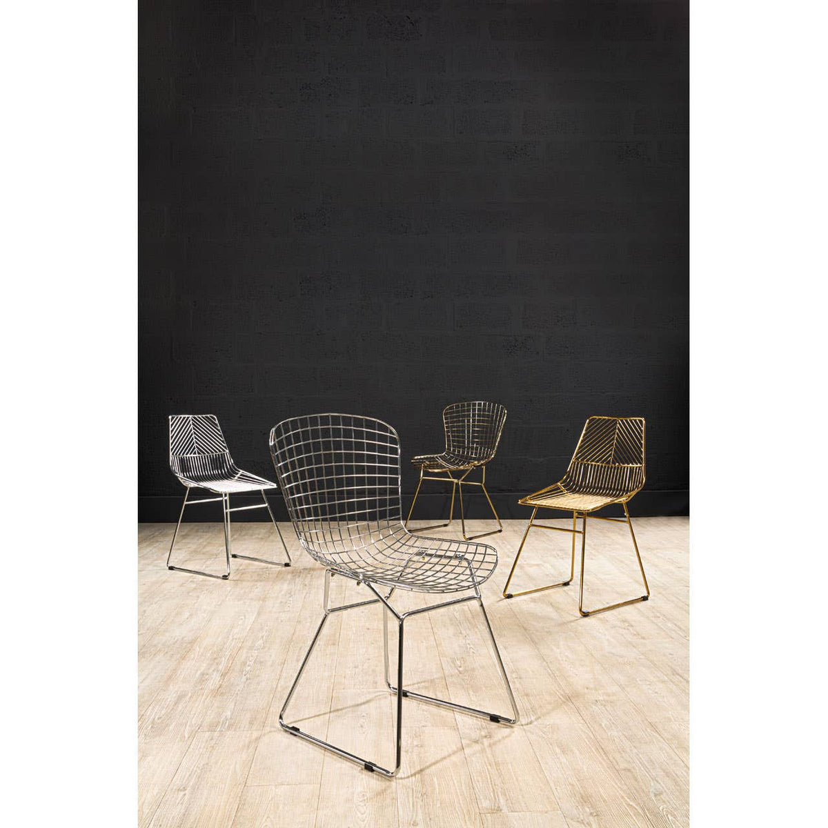 A collection of four wire chairs with metal grid frames, featuring a gold chair in the foreground and three other chairs in varying colors behind it, set against a black wall and a light wooden floor