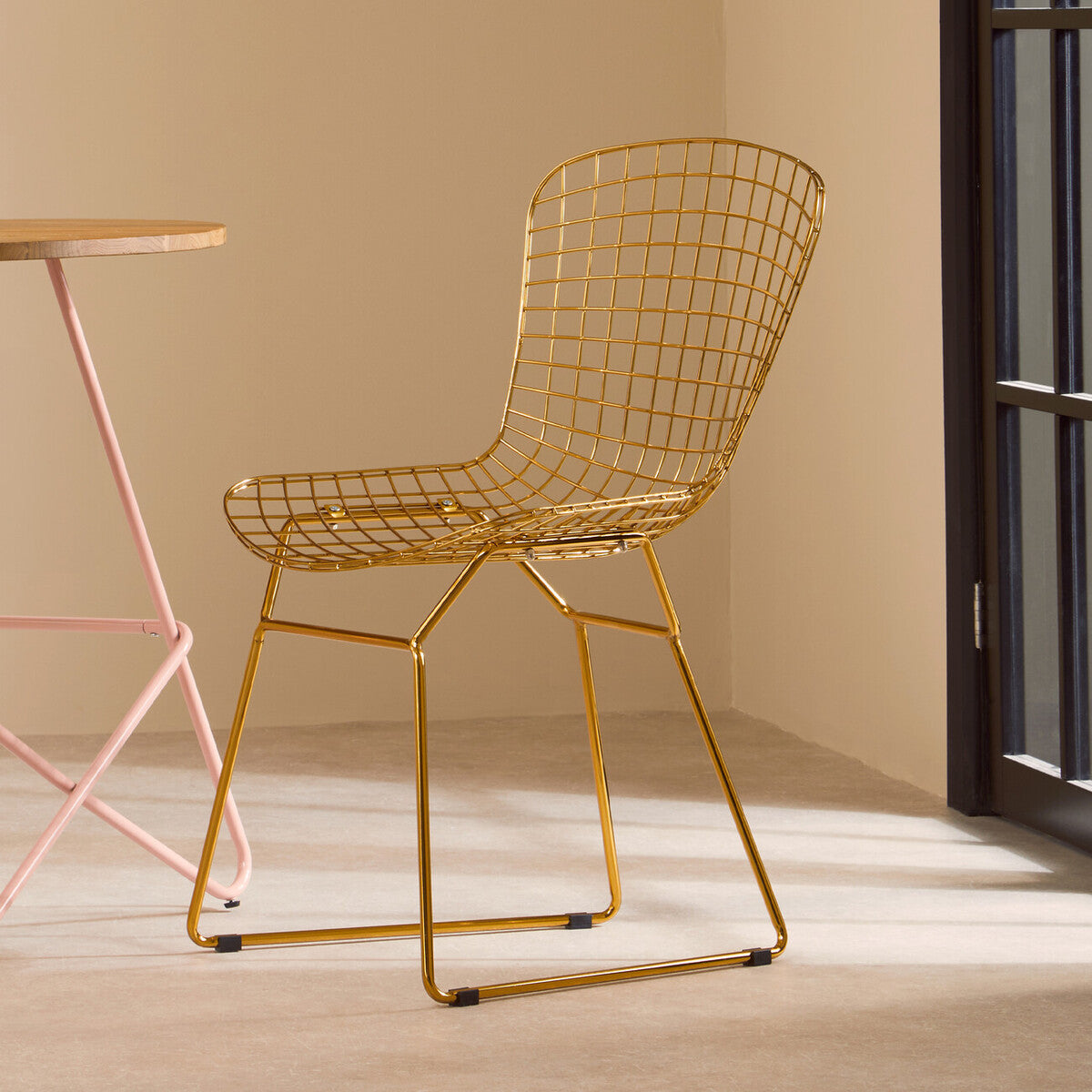 Gold metal grid frame wire chair with a modern design, featuring a curved backrest and open seat, positioned next to a pink metal table in a softly lit room with beige walls