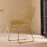 Gold metal grid frame wire chair with a modern design, featuring a curved backrest and open seat, positioned next to a pink metal table in a softly lit room with beige walls