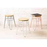 Three hairpin stools with wooden seats and metal legs in grey, copper, and black finishes are arranged against a white brick wall. The stools feature a minimalist design with slender, hairpin-style legs and round tops