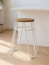 A grey metal hairpin stool with a round wooden seat, featuring a minimalist design and slender legs, placed on a light wooden floor in a bright, airy room