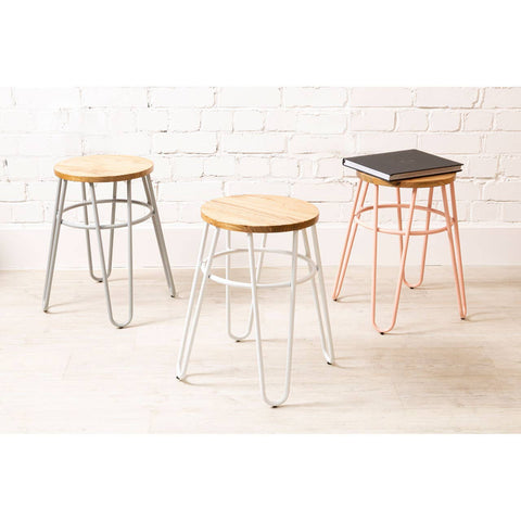 Three hairpin stools are displayed against a white brick wall. The stools feature round wooden seats made of elm wood, with one stool having a pink metal base, another in silver, and the third in black. The design is modern and minimalist, showcasing the contrast between the metal legs and the natural wood