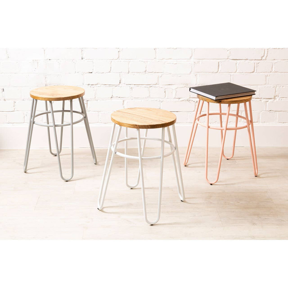 Three hairpin stools are displayed against a white brick wall. The stools feature round wooden seats made of elm wood, with one stool having a pink metal base, another in silver, and the third in black. The design is modern and minimalist, showcasing the contrast between the metal legs and the natural wood