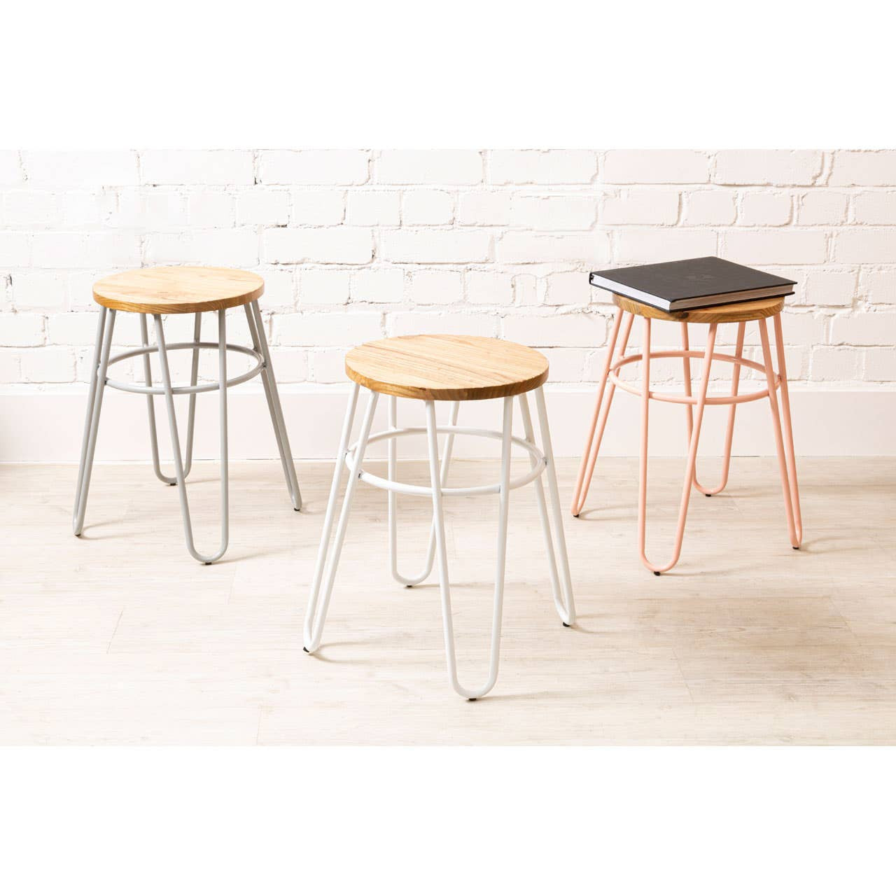 Three hairpin stools are displayed against a white brick wall. The stools feature round wooden seats made of elm wood, with one stool having a pink metal base, another in silver, and the third in black. The design is modern and minimalist, showcasing the contrast between the metal legs and the natural wood