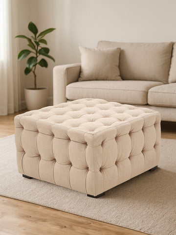 Beige linen button-tufted stool with a square shape, featuring a soft, padded top and decorative buttons, placed on a light wooden floor in a cozy living room setting. A beige sofa and a potted plant are visible in the background
