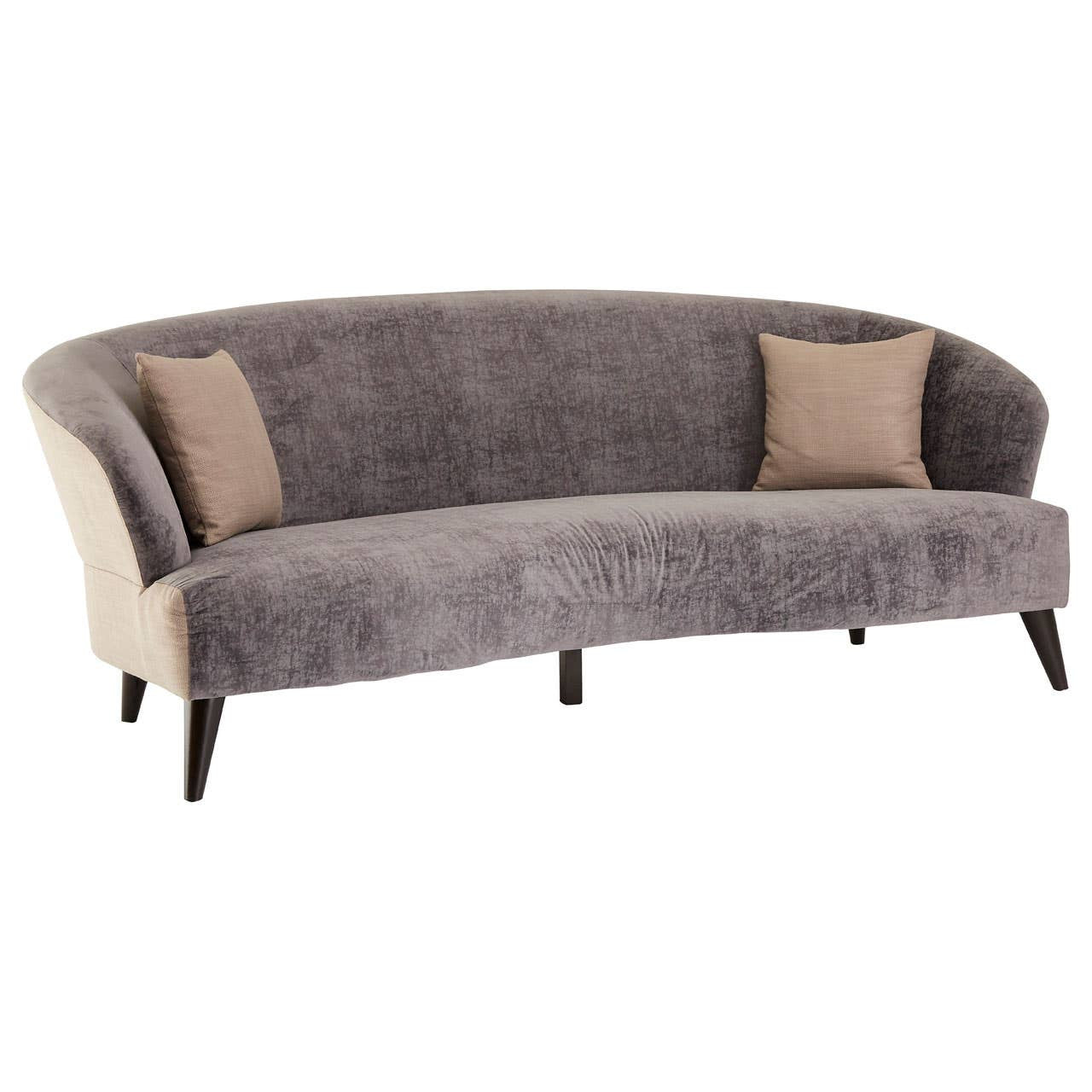 A three-seat sofa upholstered in grey velvet with a curved design, featuring two beige accent pillows on either side and sleek wooden legs