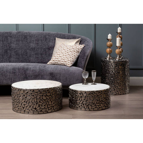 Grey velvet sofa with a curved backrest and plush cushions, accompanied by two round coffee tables with textured bases and white tops, set against a dark blue wall. A decorative pillow rests on the sofa, and a tall decorative piece with wooden accents stands beside the tables