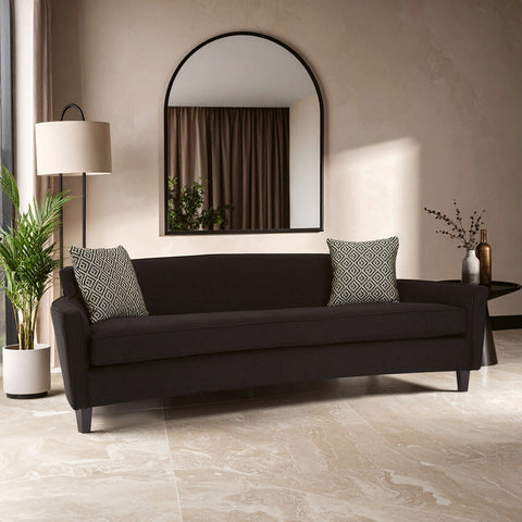 A three-seat sofa upholstered in black dimity fabric, featuring a sleek and modern design. The sofa has two light gray accent pillows and is positioned in a stylish living space with a large round mirror, a floor lamp, and a potted plant nearby. The flooring is light-colored, complementing the overall aesthetic