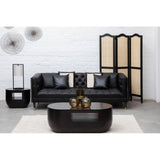 A stylish black faux leather three-seat sofa with decorative cushions, positioned in a modern living room setting. The sofa is complemented by a round coffee table and a decorative room divider in the background. The space features a light-colored rug and wall decor, creating a contemporary ambiance
