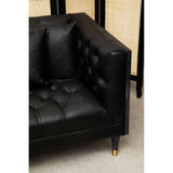 Black faux leather sofa with a tufted design, featuring two decorative pillows, set against a light-colored floor and a backdrop of a woven screen. The sofa has sleek, tapered legs with gold accents