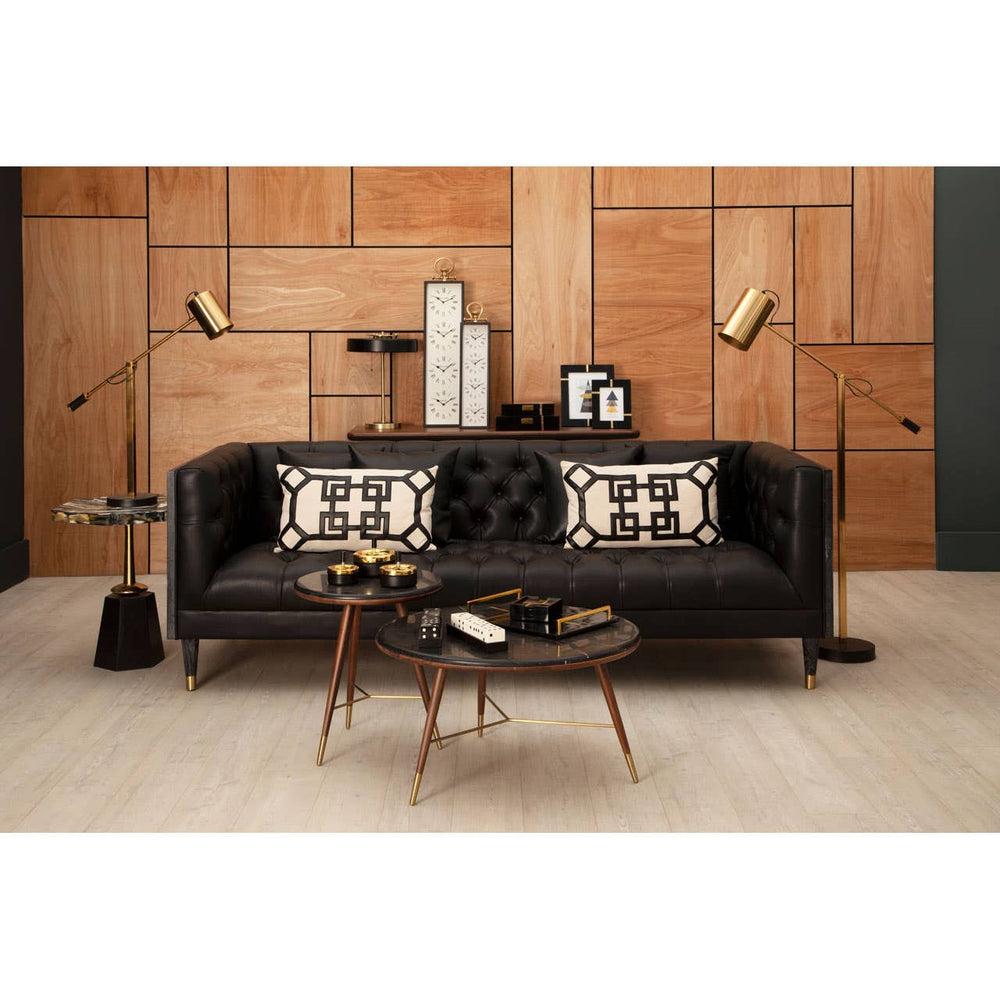 Three-seat black faux leather sofa with a modern design, featuring decorative pillows with geometric patterns. The sofa is positioned against a wooden wall, complemented by stylish floor lamps and a round coffee table with books and a decorative item on top. The flooring is light-colored wood, enhancing the contemporary aesthetic of the space