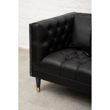 Black faux leather sofa with tufted detailing on the armrest and seat, featuring wooden legs with gold accents. The corner of the sofa is visible, showcasing its sleek design and modern aesthetic against a light-colored floor