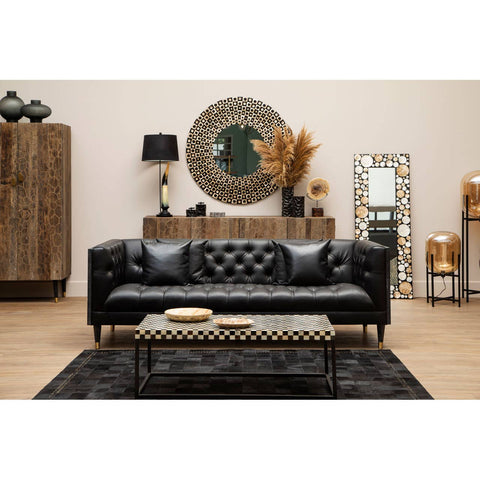 Black faux leather three-seat sofa with tufted detailing, positioned in a stylish living room setting. The sofa is complemented by a patterned black and white rug, a round decorative mirror on the wall, and wooden furniture pieces in the background, including a cabinet and a lamp. Decorative elements such as plants and vases add warmth to the space