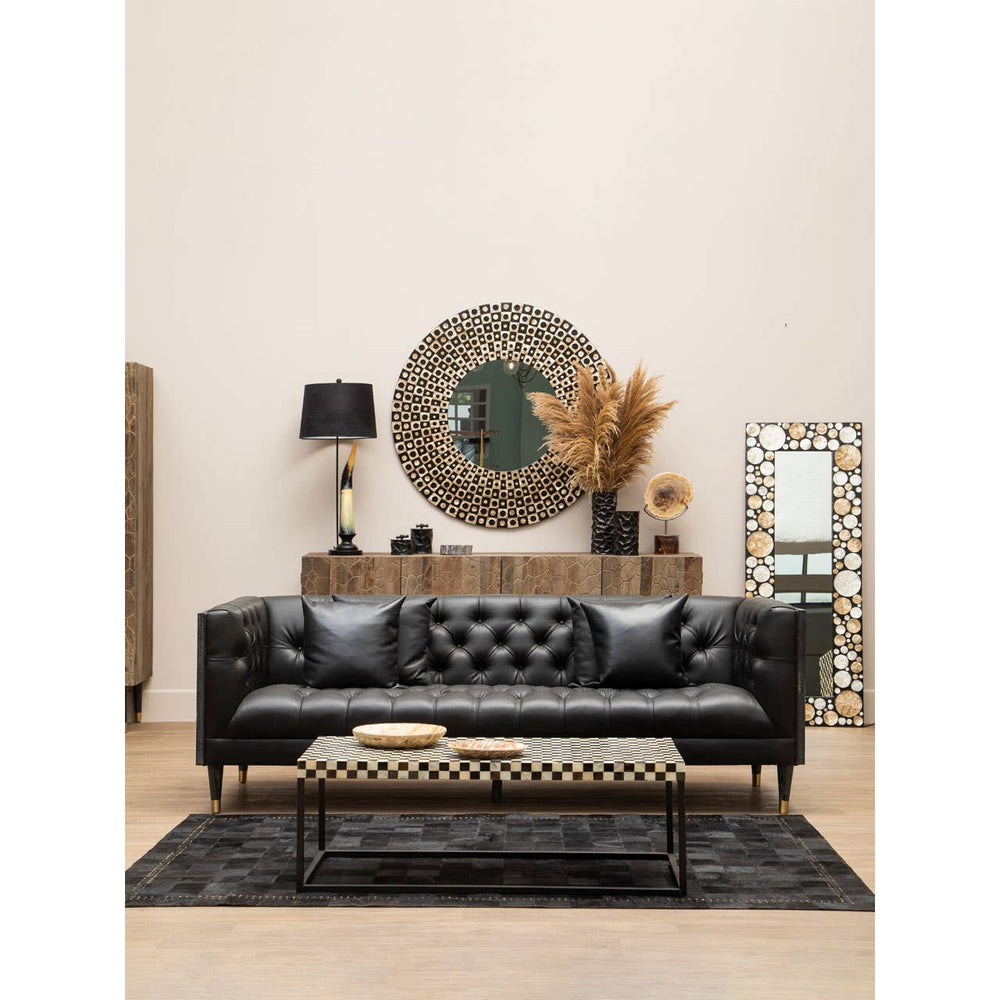 A stylish black faux leather sofa with a tufted design, featuring three seats and decorative cushions. The sofa is placed in a modern living room setting with a round mirror, a lamp, and a decorative plant in the background, along with a patterned rug underneath