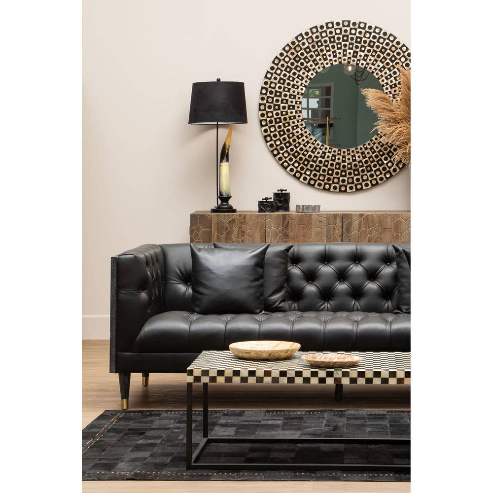 A black faux leather three-seat sofa with tufted detailing, positioned against a light-colored wall. A round decorative mirror hangs above the sofa, reflecting a stylish lamp with a black shade on a wooden side table. A patterned coffee table sits in front of the sofa, accompanied by two round wooden coasters, and a textured black rug covers the floor