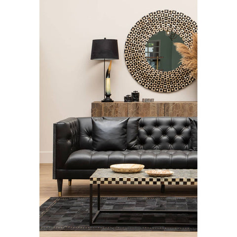A black faux leather three-seat sofa with tufted detailing, positioned against a light-colored wall. A round decorative mirror hangs above the sofa, reflecting a stylish lamp with a black shade on a wooden side table. A patterned coffee table sits in front of the sofa, accompanied by two round wooden coasters, and a textured black rug covers the floor