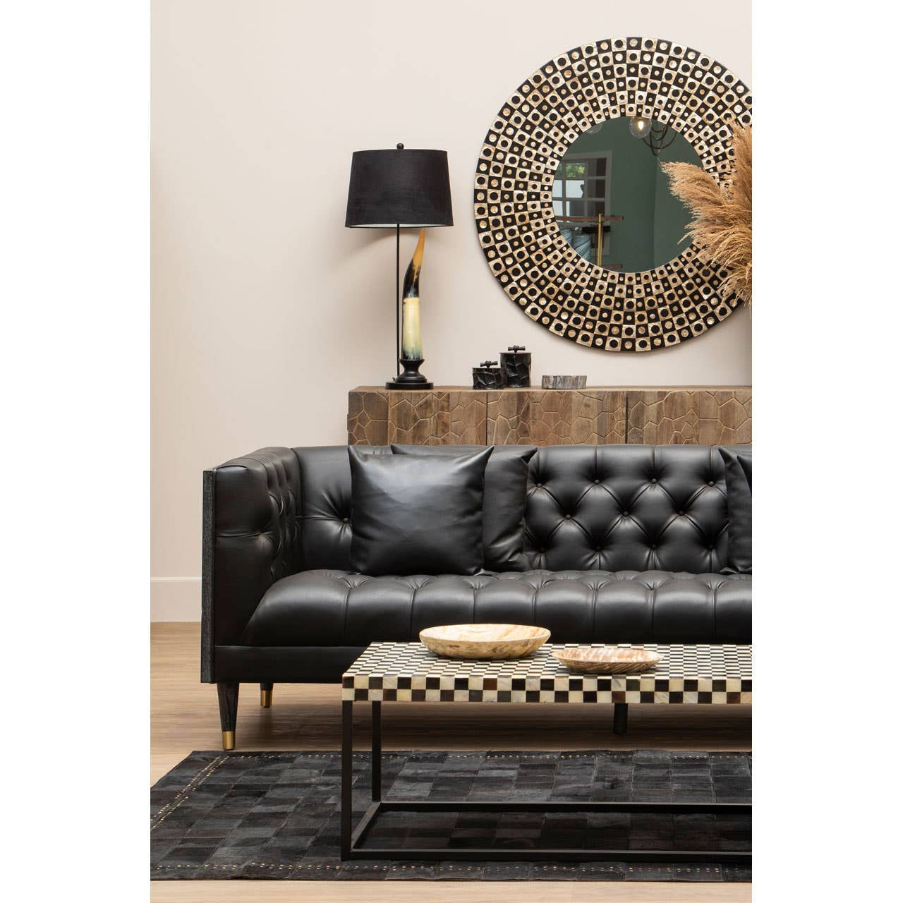 A black faux leather three-seat sofa with tufted detailing, positioned against a light-colored wall. A round decorative mirror hangs above the sofa, reflecting a stylish lamp with a black shade on a wooden side table. A patterned coffee table sits in front of the sofa, accompanied by two round wooden coasters, and a textured black rug covers the floor