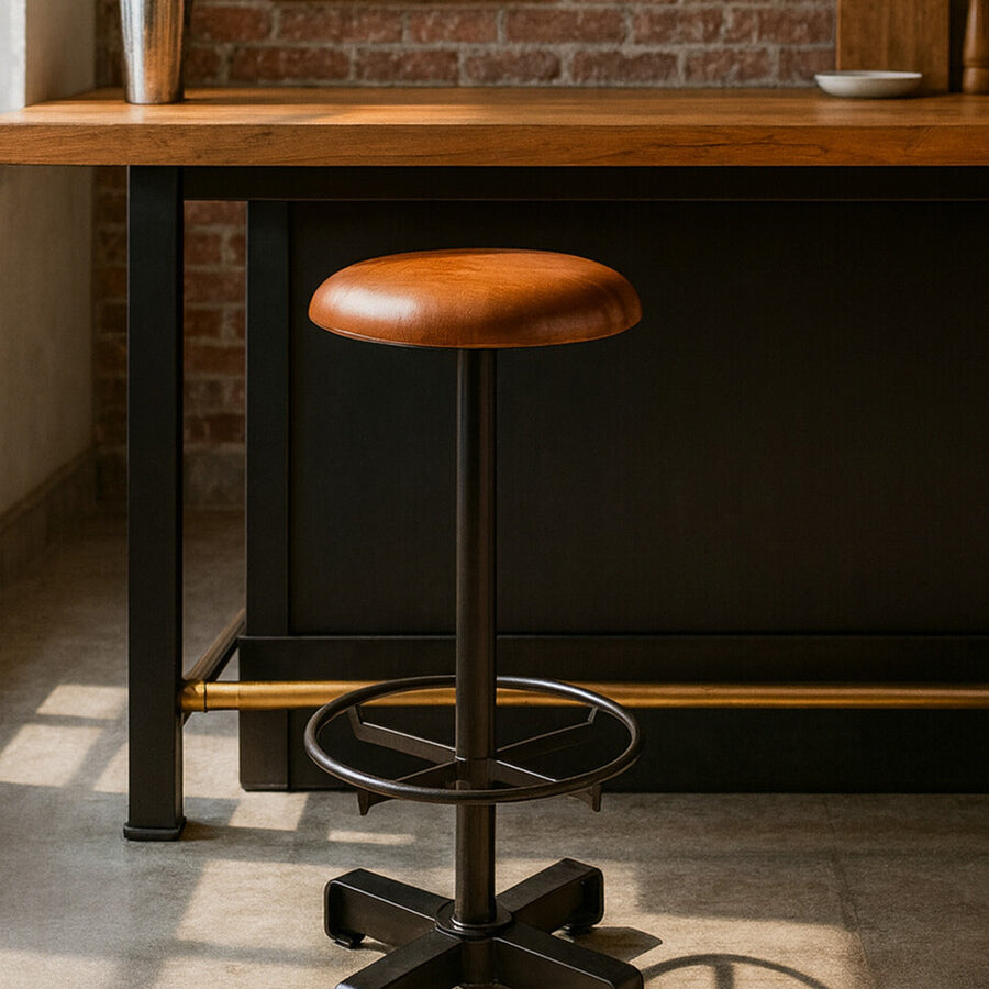 Light brown leather bar stool with a round seat and a black metal base, featuring a footrest and a sleek, modern design. The stool is positioned next to a wooden bar counter in a well-lit room with brick walls and framed artwork in the background