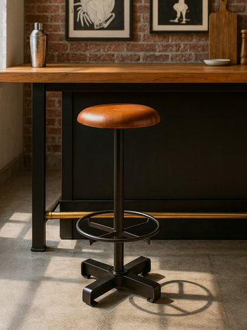 Light brown leather bar stool with a round seat and a black metal base, featuring a footrest and a sleek, modern design. The stool is positioned next to a wooden bar counter in a well-lit room with brick walls and framed artwork in the background
