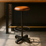 Light brown leather bar stool with a round seat and a black metal base featuring a circular footrest and a star-shaped base, positioned on a light-colored floor near a wooden countertop