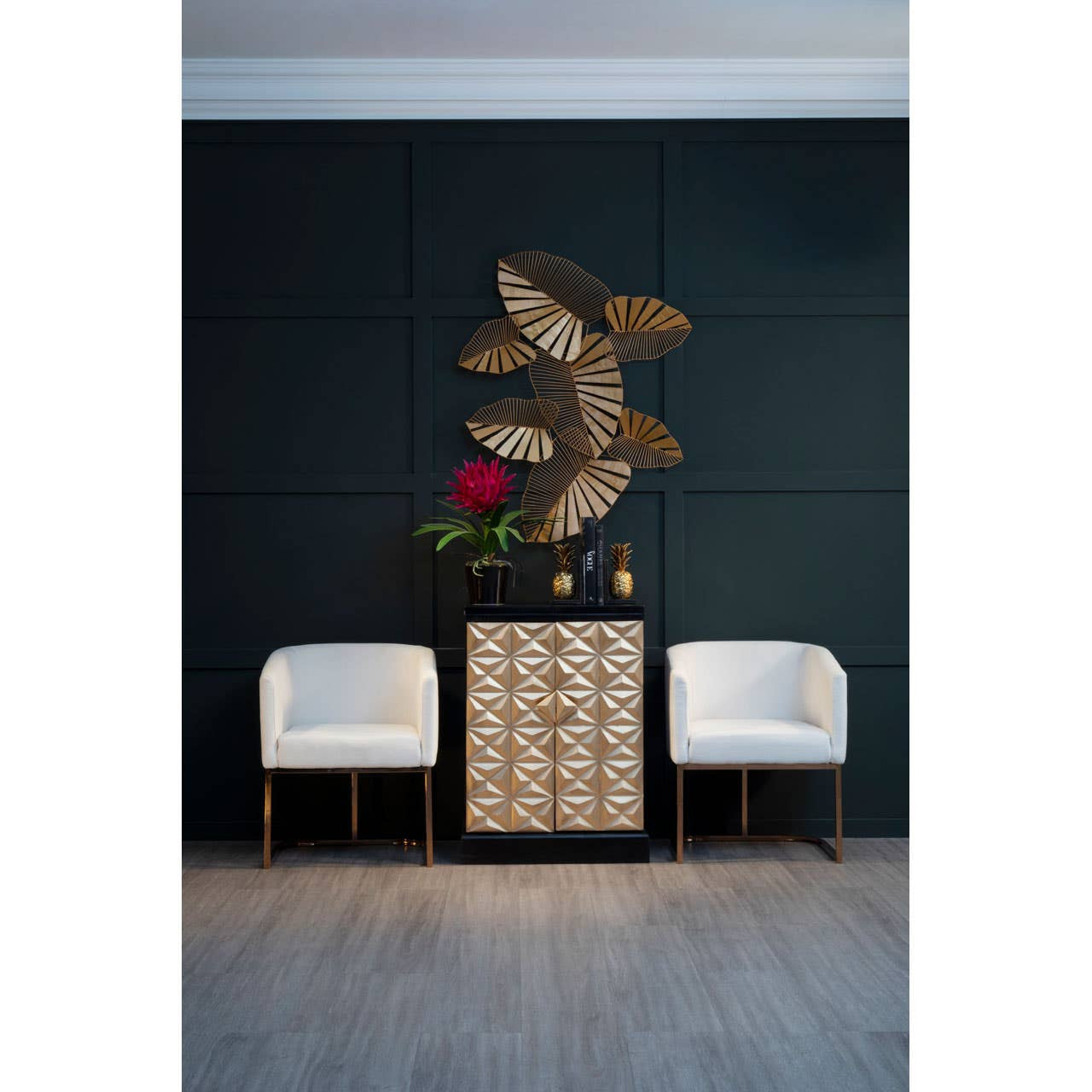 A stylish bar cabinet made of mango wood featuring a gold and black finish, positioned against a dark green wall. The cabinet has multiple drawers with a sleek design, complemented by two modern white chairs on either side. Above the cabinet, there is a decorative wall art piece with a floral design. The floor is a light wood finish, enhancing the contemporary aesthetic of the space