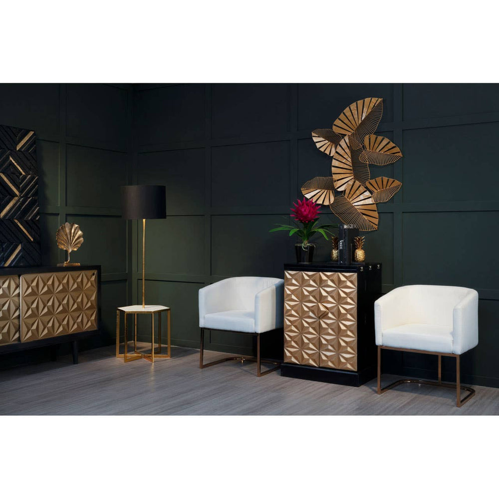 A stylish bar cabinet featuring a gold and black design, made from mango wood, positioned against a dark green wall. The cabinet has a geometric pattern on its front and is complemented by two white upholstered chairs with gold legs. A decorative wall piece and a small side table are also visible in the scene, along with a vase of red flowers on top of the cabinet
