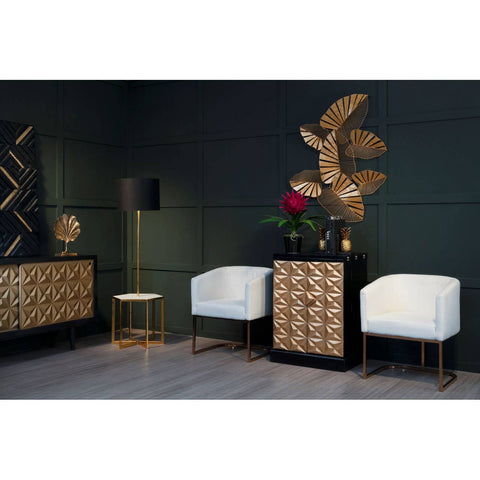 A stylish bar cabinet featuring a gold and black design, made from mango wood, positioned against a dark green wall. The cabinet has a geometric pattern on its front and is complemented by two white upholstered chairs with gold legs. A decorative wall piece and a small side table are also visible in the scene, along with a vase of red flowers on top of the cabinet