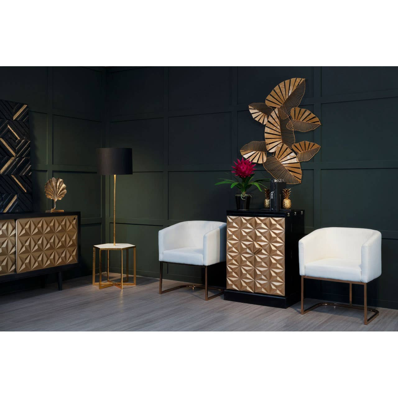 A stylish bar cabinet featuring a gold and black design, made from mango wood, positioned against a dark green wall. The cabinet has a geometric pattern on its front and is complemented by two white upholstered chairs with gold legs. A decorative wall piece and a small side table are also visible in the scene, along with a vase of red flowers on top of the cabinet