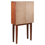 A tall cabinet made from mango wood featuring a warm brown finish, with two textured doors displaying a geometric pattern. The cabinet stands on four slender legs, adding a modern touch to its coastal design