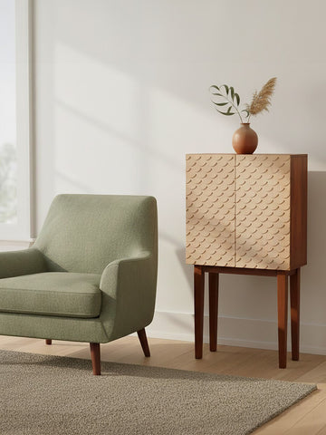 A coastal carved mango wood cabinet with a textured front, standing on four wooden legs, next to a light green armchair. A decorative vase with a plant is placed on top of the cabinet, and soft natural light filters through a nearby window, casting shadows on the floor