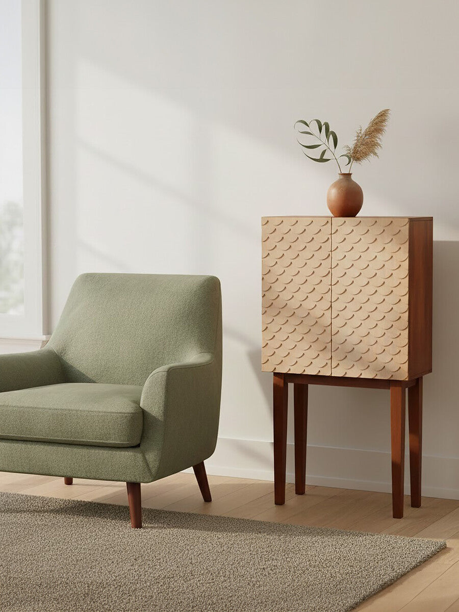 A coastal carved mango wood cabinet with a textured front, standing on four wooden legs, next to a light green armchair. A decorative vase with a plant is placed on top of the cabinet, and soft natural light filters through a nearby window, casting shadows on the floor