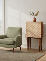 A coastal carved mango wood cabinet with a textured front, standing on four wooden legs, next to a light green armchair. A decorative vase with a plant is placed on top of the cabinet, and soft natural light filters through a nearby window, casting shadows on the floor