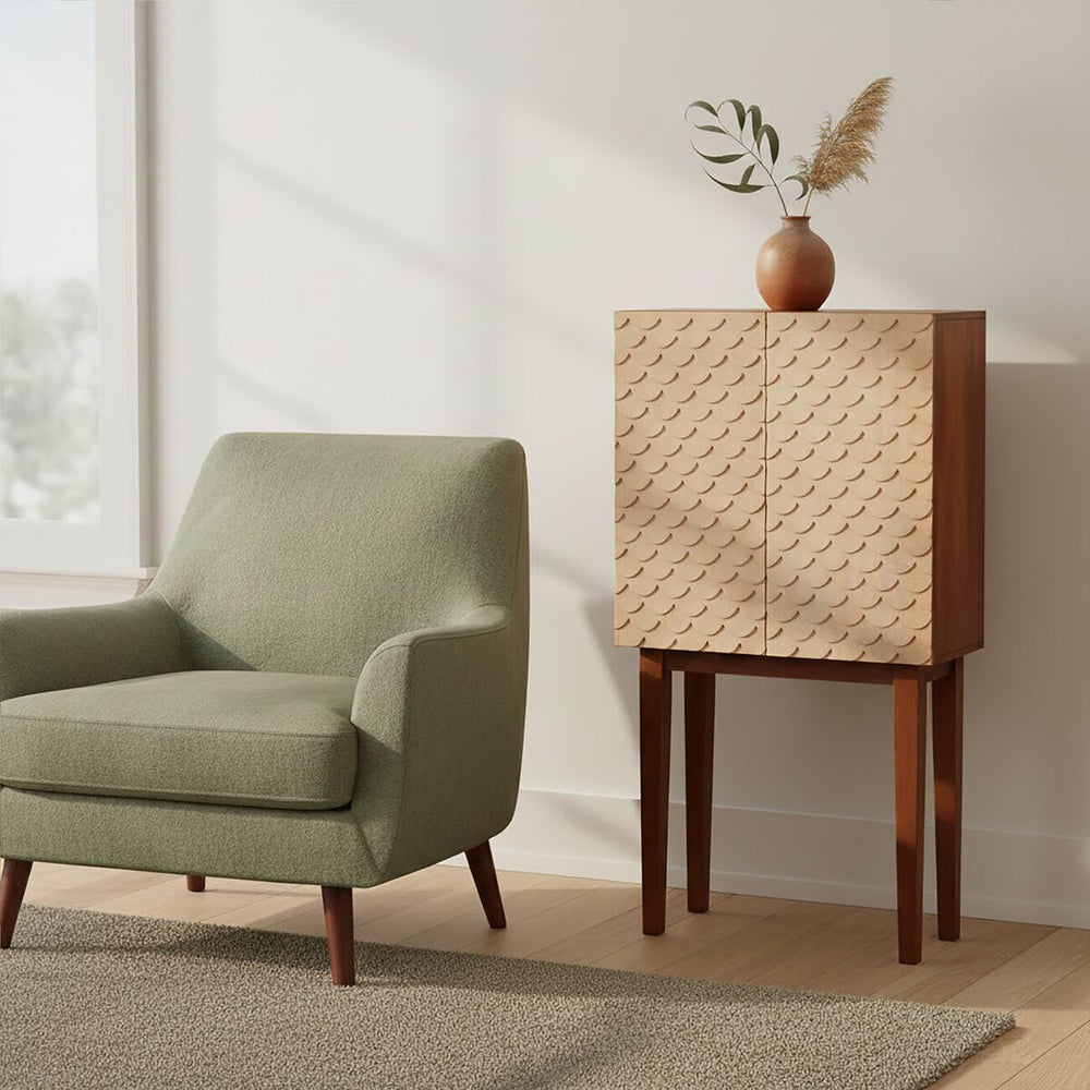 A coastal carved mango wood cabinet with a textured front design, standing on four wooden legs, next to a light green upholstered armchair. The cabinet is positioned against a wall with natural light streaming in from a nearby window, and a decorative vase with dried flowers sits on top of the cabinet. A neutral-toned rug is visible on the floor