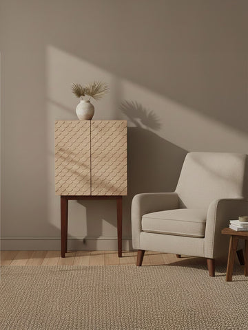 Coastal carved mango wood cabinet with a light wood finish, featuring two doors with a subtle wave pattern, positioned beside a light gray upholstered armchair and a small wooden side table. A simple white vase with a single flower sits atop the cabinet, against a neutral wall and soft beige carpet