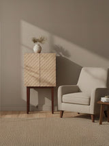 Coastal carved mango wood cabinet with a light wood finish, featuring two doors with a subtle wave pattern, positioned beside a light gray upholstered armchair and a small wooden side table. A simple white vase with a single flower sits atop the cabinet, against a neutral wall and soft beige carpet