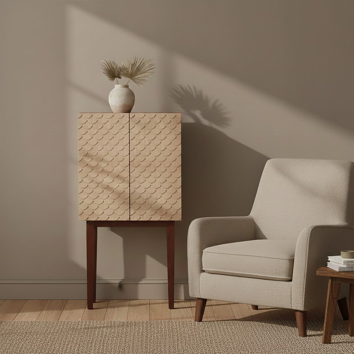 A coastal carved mango wood cabinet with a textured front design, standing on four wooden legs, next to a light-colored upholstered armchair. A simple white vase with dried flowers sits atop the cabinet, and soft shadows create a warm atmosphere in the room