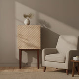 A coastal carved mango wood cabinet with a textured front design, standing on four wooden legs, next to a light-colored upholstered armchair. A simple white vase with dried flowers sits atop the cabinet, and soft shadows create a warm atmosphere in the room