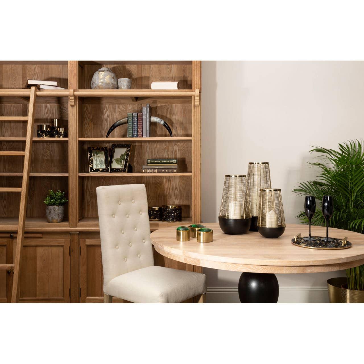 Round wooden dining table with beige upholstered chair, gold vases, and bookshelf backdrop